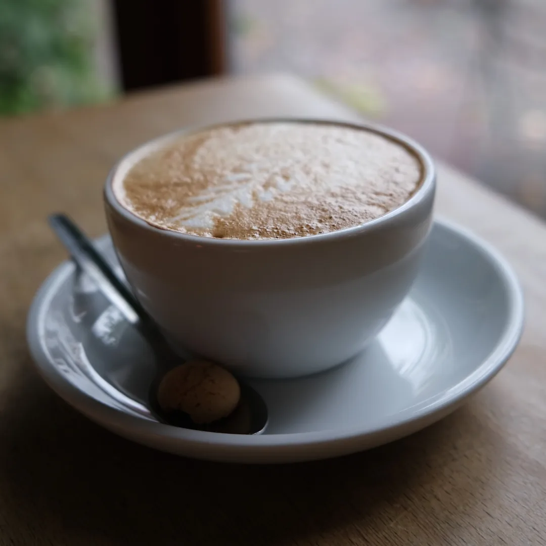 A real cappuccino in a white cup and saucer with a spoon and biscuit on a wooden table.