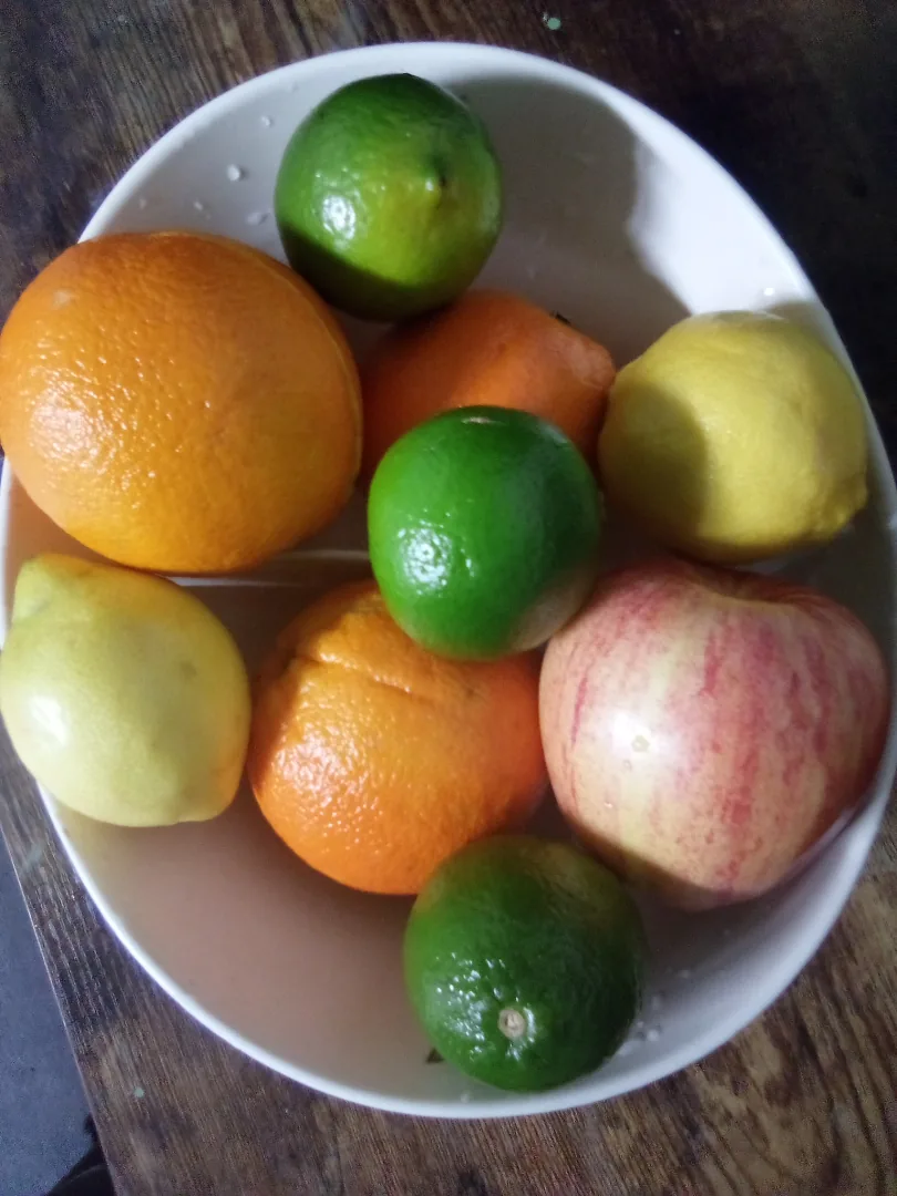 A real white oval bowl holding oranges, limes and apples on a wooden surface.