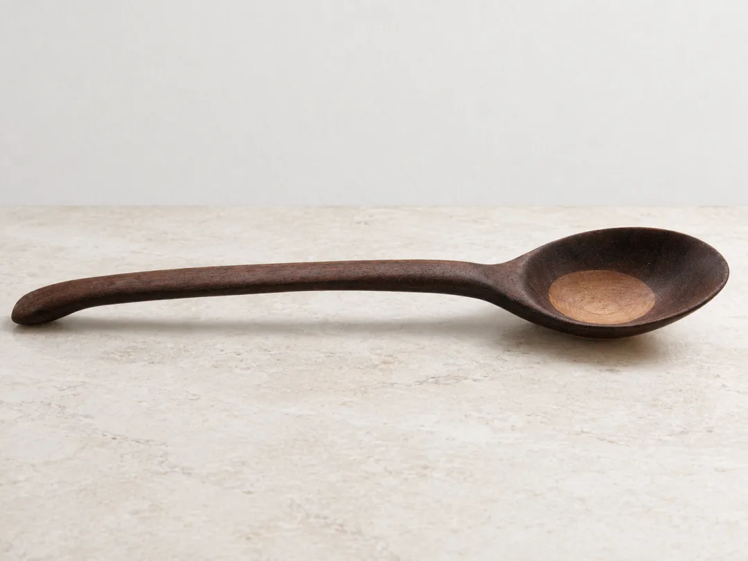 A generated product scene showing the same recognizable wooden spoon on pale stone against a neutral wall, with softer natural light and a contact shadow.